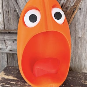 May include: An orange pumpkin-shaped container with a wide, open mouth and a protruding tongue. The figure features two large white eyes with black pupils and a small green stem on top. The surface is smooth and bright orange in color.