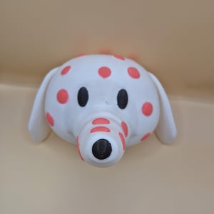 May include: A white elephant figurine with red polka dots. The elephant has black eyes and a black textured trunk. The ears are white and the background is a light tan color.