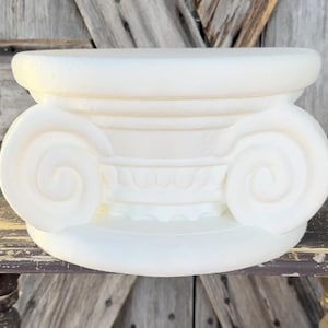 May include: A white, decorative Ionic column capital. The capital features a scrolled volute design on each side, with a detailed frieze below the top. The object is made of a solid, opaque material and sits on a wooden surface.