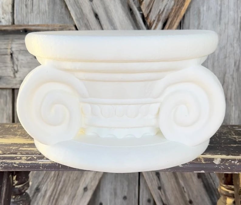May include: A white, decorative Ionic column capital. The capital features a scrolled volute design on each side, with a detailed frieze below the top. The object is made of a solid, opaque material and sits on a wooden surface.