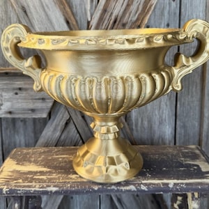 May include: A gold-colored decorative urn with ornate handles and a detailed, textured surface. The urn has a wide, open top and sits on a pedestal base. The background is a weathered wooden surface.
