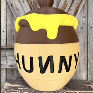 May include: A round, light tan ceramic jar with a dark brown lid featuring a yellow, dripping honey design. The word HUNNY is printed in large, bold, black capital letters across the front of the jar, which has a smooth, matte finish.
