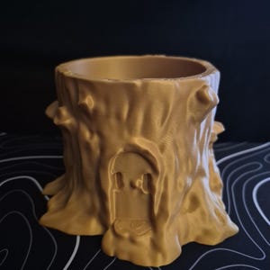 May include: A gold-colored, tree stump-shaped planter with a door design. The planter has a textured surface resembling tree bark, with protruding knots. The door is a recessed design. The planter is suitable for small plants or decorative items.