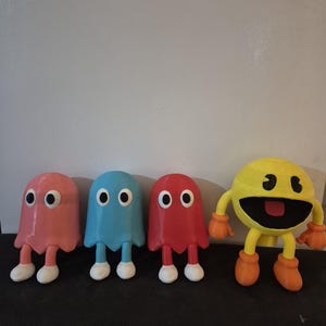 May include: Four colorful Pac-Man character figures. Three ghost figures are pink, blue, and red, each with white feet and large black eyes. A yellow Pac-Man figure with orange gloves and feet completes the set.