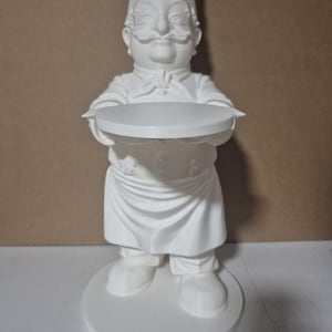 May include: A white ceramic chef figurine, holding a round tray. The chef is wearing a tall hat, a bow tie, and an apron. The figurine is standing on a circular base. The overall design is detailed and decorative.
