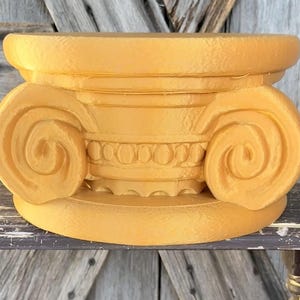 May include: A golden-yellow, decorative column capital with intricate detailing. The capital features a scrolled volute design on each side, with a detailed band of circular and linear elements. The piece is made of a solid material and sits on a flat base.