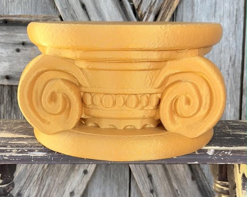 May include: A golden-yellow, decorative column capital with intricate detailing. The capital features a scrolled volute design on each side, with a detailed band of circular and linear elements. The piece is made of a solid material and sits on a flat base.