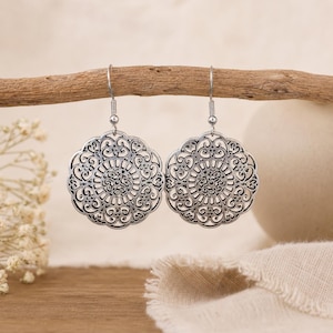 May include: A pair of silver-toned dangle earrings featuring a circular, openwork filigree design with intricate scrollwork patterns. The earrings are suspended from simple silver-toned hooks, showcasing a detailed, symmetrical metal cutout motif.