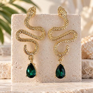 May include: A pair of gold-toned snake-shaped earrings with a textured, scale-like finish. Each earring features a curved, serpentine body that leads to a dangling, pear-shaped emerald green gemstone set in a gold-toned metal frame.