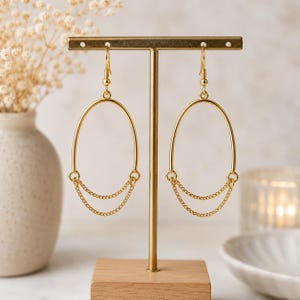 May include: Gold-toned oval hoop earrings with delicate chain accents. The earrings are displayed on a gold stand with a wooden base. The design is minimalist and elegant, suitable for everyday wear or special occasions.
