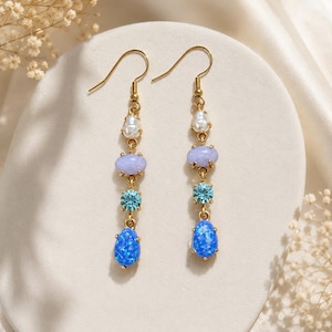 May include: A pair of gold-toned dangle earrings. Each earring features a pearly white bead, a light purple oval stone, a light blue crystal, and a blue opal-like teardrop. The earrings are displayed on a white surface.