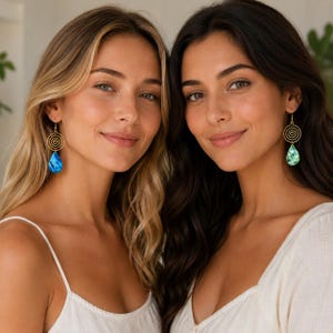 Op de afbeelding: Twee vrouwen dragen gouden spiraalvormige oorbellen met druppelvormige hangers. Eén oorbel heeft een blauwe hanger, de andere een groene hanger. Beide vrouwen dragen witte tops en lachen in de camera.