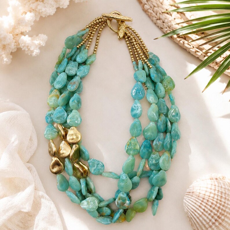 May include: A multi-strand necklace featuring polished turquoise-colored teardrop beads and several smooth gold-toned metal accents. The strands are connected by a gold-toned toggle clasp, creating a layered design with a mix of blue and gold.