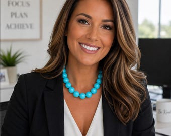 May include: A bright turquoise beaded necklace featuring large, uniform spherical beads strung together. The necklace is worn around the neck, resting against a white top layered under a black blazer, creating a bold contrast with the vibrant blue.