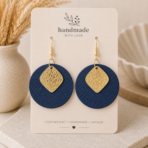 May include: A pair of dangle earrings featuring a navy blue circular leather piece with a textured gold diamond-shaped charm attached. The earrings are displayed on a cream-colored card with the text handmade WITH LOVE and LIGHTWEIGHT HANDMADE UNIQUE.