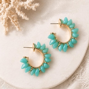 May include: A pair of gold-tone hoop earrings with turquoise teardrop-shaped beads. The earrings are arranged in a semi-circle around the gold hoops. The earrings are displayed on a white surface with a coral branch in the background.