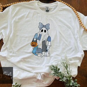 Puede incluir: Una camiseta blanca de manga corta con el gráfico de un fantasma que lleva un lazo de cuadros azules y sostiene un cubo de calabaza naranja. El fantasma está decorado con varios patrones de patchwork azules y blancos por todo el cuerpo.