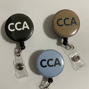 May include: Three retractable badge holders with round buttons that say "CCA" in white letters on a black background, black letters on a brown background, and black letters on a light blue background.