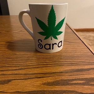 May include: White ceramic mug with a green marijuana leaf design and the name "Sara" written in black lettering.