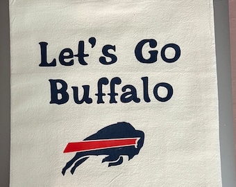 May include: White kitchen towel with the text "Let's Go Buffalo" in navy blue. Below the text is a navy blue buffalo logo with a red stripe. The towel is folded.