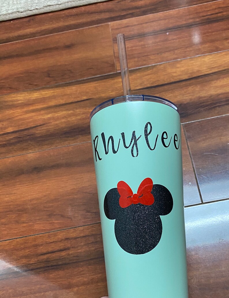 Może przedstawiać: Zielony, wielokrotnego użytku kubek z nadrukiem czarnej głowy Myszki Miki z brokatem i imieniem "Rhylee" napisanym czarnym, ozdobnym pismem. Kubek ma przezroczystą słomkę z tworzywa sztucznego.