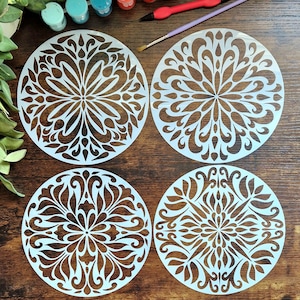 May include: Four circular stencils featuring intricate, symmetrical mandala-style patterns with cut-out floral and leaf-like shapes. The stencils are translucent white and arranged in a two-by-two grid, accompanied by small paint pots and brushes.