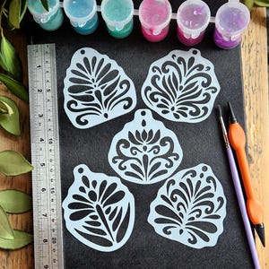 May include: A collection of light blue stencils with floral designs, arranged on a black surface. Paint pots in various colors, including blue, pink, and purple, are visible at the top. Paint brushes and a ruler are also present.