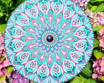 May include: A round, hand-painted mandala in shades of teal, pink, and purple, with a central black bead. The intricate design features a symmetrical pattern of floral and geometric shapes. The artwork is held against a backdrop of green foliage and pink flowers.
