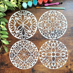 May include: Four white, circular stencils with intricate floral and geometric patterns are arranged on a wooden surface. Small paint pots in various colors, along with paintbrushes and tools, are visible. Green foliage is in the background.