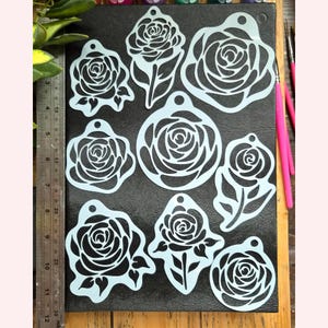 May include: A collection of eight light blue rose-shaped stencils with circular hanging holes. A metal ruler is positioned on the left side, showing the largest stencil is approximately 4 inches wide. The stencils feature various rose designs.