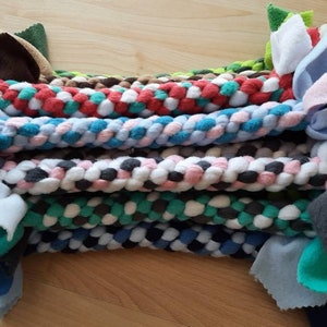 Könnte beinhalten: Ein Stapel bunter geflochtener Hundespielzeuge mit verknoteten Fleece-Enden. Die Spielzeuge sind aus verschiedenen Farben von Fleece-Stoff gefertigt, darunter Rot, Blau, Grün, Weiß und Schwarz.