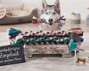 May include: A braided fleece tug toy for dogs featuring a multi-colored pattern of pink, blue, white, and dark grey. The toy has knotted ends and is displayed alongside a slate sign with the text Handgefertigtes Zergelseil aus weichem Fleece.