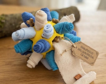 May include: A yellow spherical dog toy featuring multiple rolled fabric strips in shades of blue, light blue, and cream. Small bone-shaped treats are tucked between the fabric rolls. A brown paper tag attached with twine reads HANDGEFERTIGTES INTELLIGENZSPIELZEUG FÜR HUNDE.