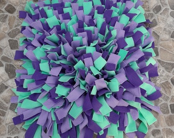 May include: A purple and green dog bed made of soft, fluffy fabric. The bed is made of many strips of fabric that are tied together.