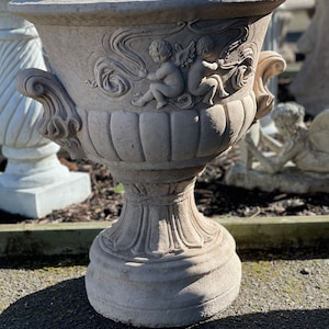 May include: A large, grey concrete planter with a decorative design of two cherubs holding a ribbon. The planter has a wide, round base and a tall, slender body.