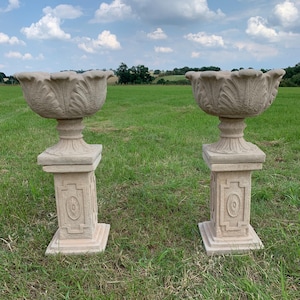 May include: Two beige stone planters with decorative, leaf-shaped detailing, mounted on pedestals with square bases. The planters are positioned in a grassy field under a cloudy sky, suitable for a garden or landscape.