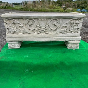 May include: A rectangular, light-coloured stone planter with ornate floral carvings. The planter sits on four short, decorative feet. The design includes detailed leaf and scroll patterns, and a central vase motif. The background shows a green surface and a rural landscape.