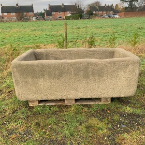 May include: A large, rectangular, grey stone trough or planter sits on a wooden pallet in a grassy field. The trough has a rough, textured surface and appears to be made of concrete or stone. The background shows a row of houses in the distance.
