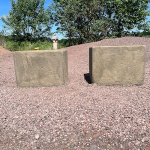 May include: Two rectangular, light-coloured concrete blocks. The blocks are set on a bed of small, reddish-brown gravel. Trees and a blue sky are visible in the background. The blocks appear to be part of a landscaping project.