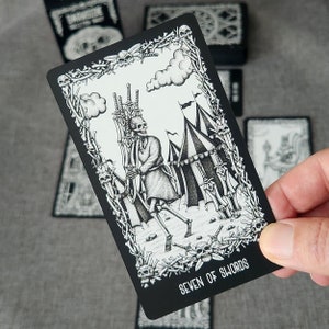 May include: Black and white tarot card illustration of the Seven of Swords. A skeletal figure is running away with seven swords, leaving behind a tent and other swords.