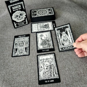 May include: A black and white tarot deck with the title "Darkside Skeleton Tarot" on the box. The cards feature illustrations of skeletons and other gothic imagery.