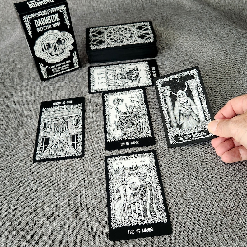 May include: A black and white tarot deck with the title "Darkside Skeleton Tarot" on the box. The cards feature illustrations of skeletons and other gothic imagery.