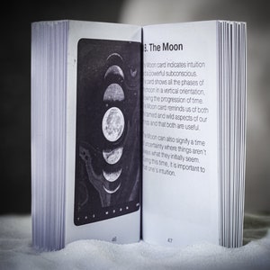 May include: A book open to a page with a black and white illustration of the moon in different phases. The text on the page reads "8. The Moon" and describes the meaning of the Moon card in a tarot deck.