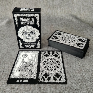 May include: A black box with white text that reads "Darkside Skeleton Tarot" and a black and white illustration of a skull. The box is sitting on a gray surface next to a stack of black tarot cards with white illustrations.