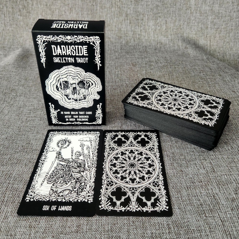 May include: A black box with white text that reads "Darkside Skeleton Tarot" and a black and white illustration of a skull. The box is sitting on a gray surface next to a stack of black tarot cards with white illustrations.