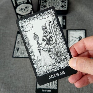 May include: Black and white tarot card with a skeletal queen sitting on a throne. The card is bordered with a design of skulls and bones. The text "QUEEN OF CUPS" is printed at the bottom of the card.