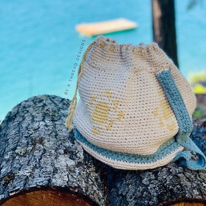 May include: A crocheted drawstring bag in cream and blue, resting on a log. The bag features a yellow design and a blue strap. The text "BY A THREAD DESIGNS" is visible on the side. The background shows a lake and a tree.