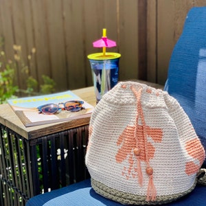May include: A white crocheted drawstring bag with a pink floral design and a brown tassel. The bag is sitting on a blue chair with a wooden table and a magazine in the background.