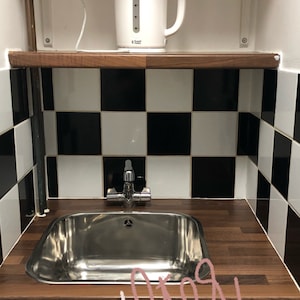 May include: A white electric kettle sits on a wooden shelf above a stainless steel sink with a chrome tap. The sink is set into a dark wood worktop. The backsplash is black and white chequered tiles.