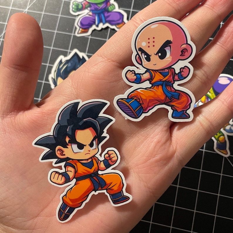 May include: A hand holding several cartoon stickers featuring characters in orange and blue outfits. The stickers are of various characters with black hair and bald heads. The stickers are on a black and gray cutting mat.